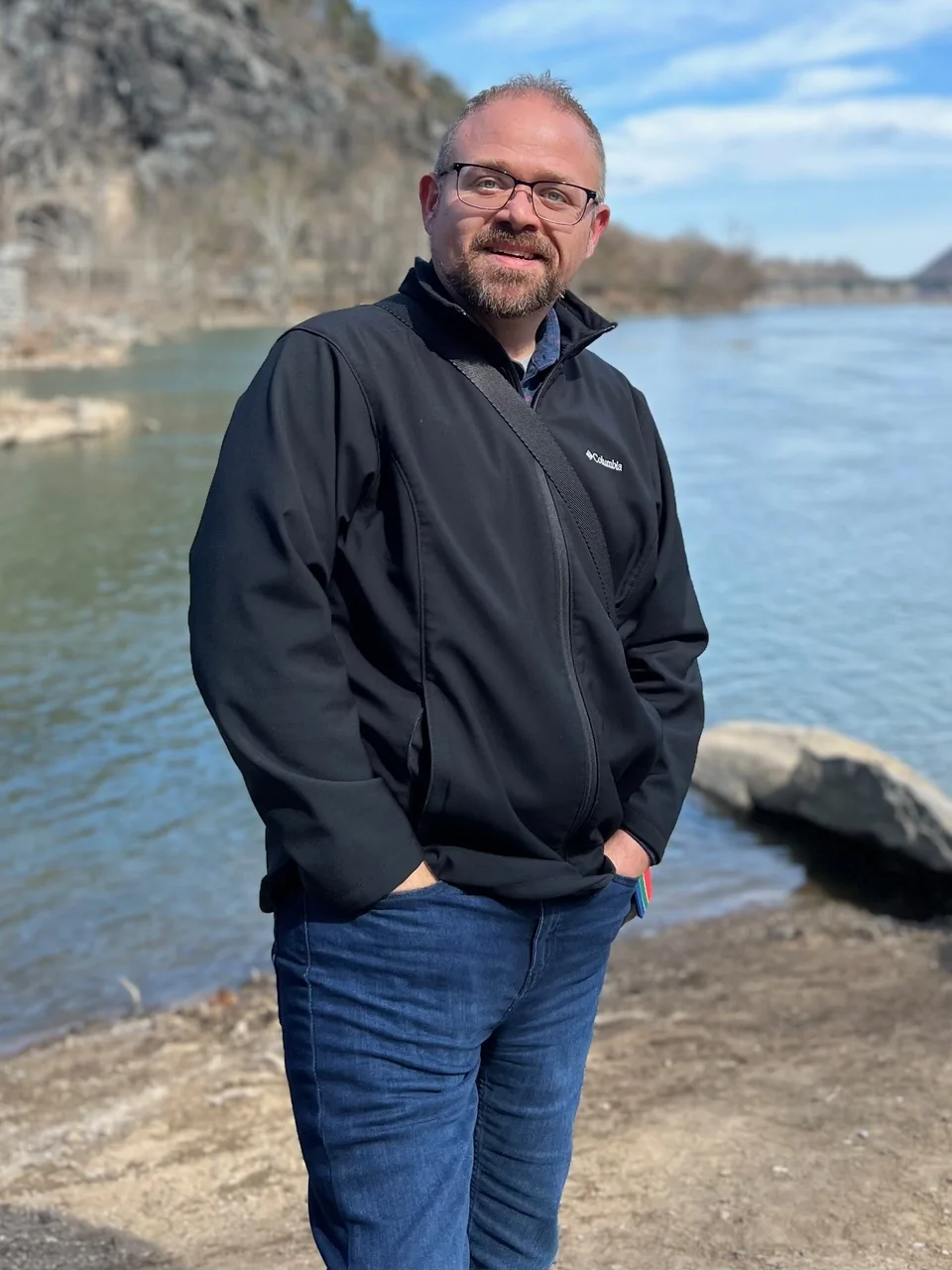 Noah Kaye standing by a river on a clear day, hands in his pockets